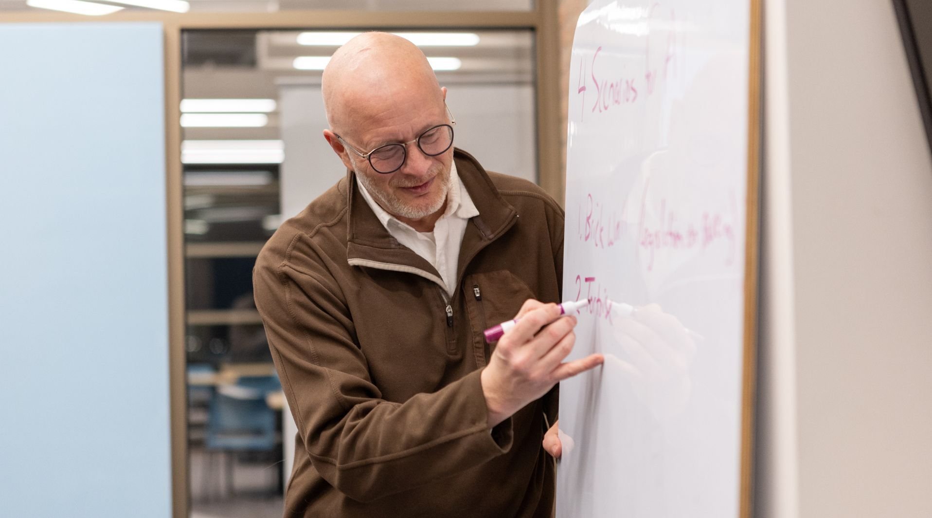 A man wearing glasses and a brown jacket writes on a whiteboard with a pink marker in a modern office setting.