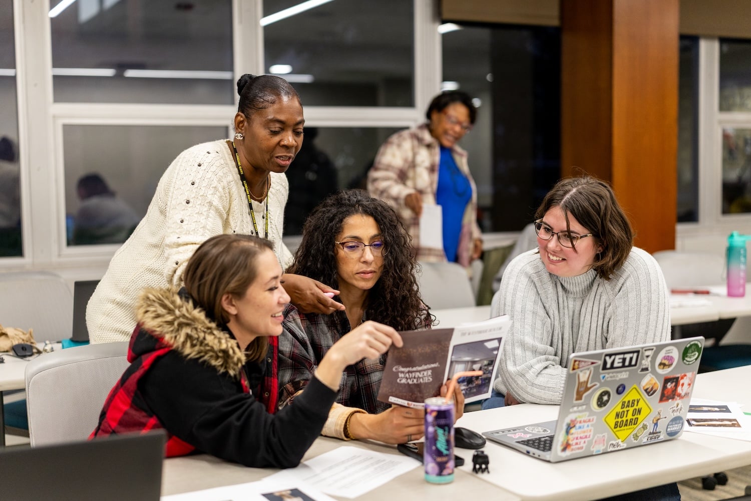 Four women sit and stand around a table in a classroom, looking at a laptop and papers, with another woman standing and smiling in the background.